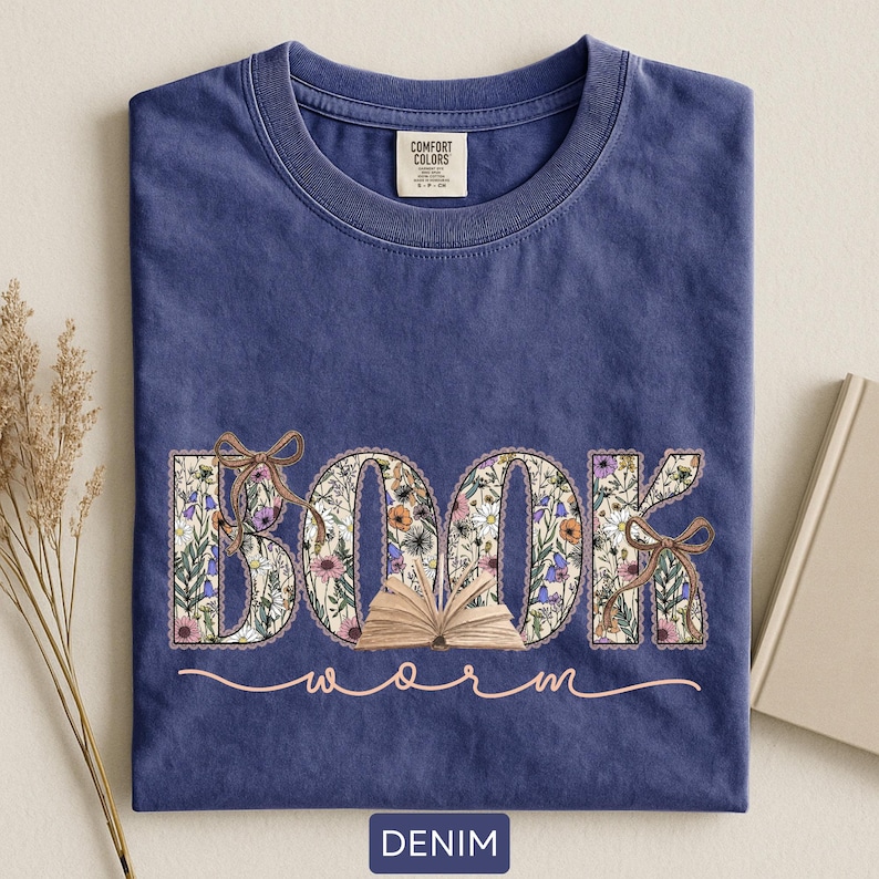 May include: A denim blue t-shirt with the word "BOOK" in floral print, an open book graphic, and the word "worm" in a cursive font. The shirt has a crew neck and a tag that says "Comfort Colors".