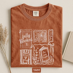 May include: A terracotta-coloured t-shirt with a white graphic design featuring a coffee cup, a library book card, and an open book with the words "One more chapter." The shirt also has the text "fully booked."