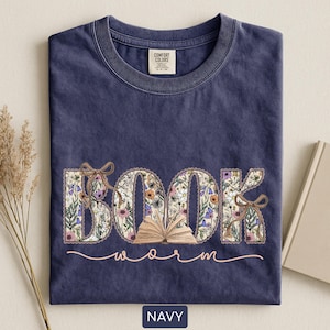 May include: Navy blue t-shirt with the word "BOOK" in floral print and an open book graphic. The word "worm" is written in a cursive font below the word "BOOK". The shirt has a crew neck and a tag that says "Comfort Colors".