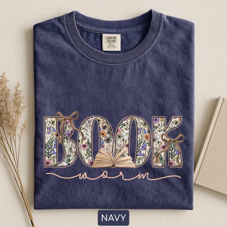 May include: Navy blue t-shirt with the word "BOOK" in floral print and an open book graphic. The word "worm" is written in a cursive font below the word "BOOK". The shirt has a crew neck and a tag that says "Comfort Colors".