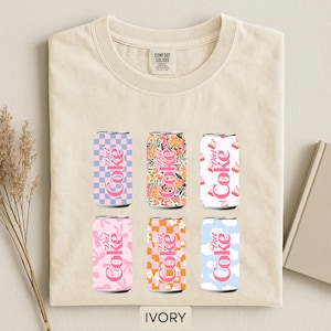 May include: Ivory-colored t-shirt featuring six Diet Coke can designs. The cans showcase various patterns, including checkered, floral, and cloud motifs, in pink, orange, and blue hues. The text "Diet Coke" is displayed on each can. The shirt has a Comfort Colors tag.