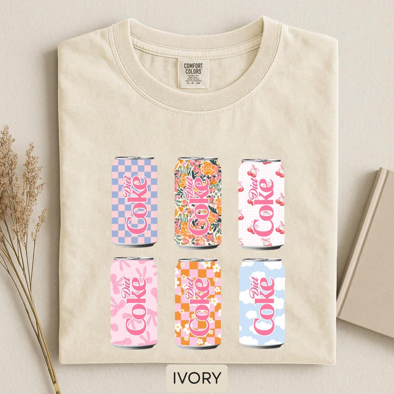 May include: Ivory-colored t-shirt featuring six Diet Coke can designs. The cans showcase various patterns, including checkered, floral, and cloud motifs, in pink, orange, and blue hues. The text "Diet Coke" is displayed on each can. The shirt has a Comfort Colors tag.