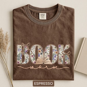 May include: Espresso-colored t-shirt with the words "BOOK worm" in a floral design. The word "BOOK" is filled with colorful flowers and an open book graphic. The shirt has a crew neck and a Comfort Colors tag.