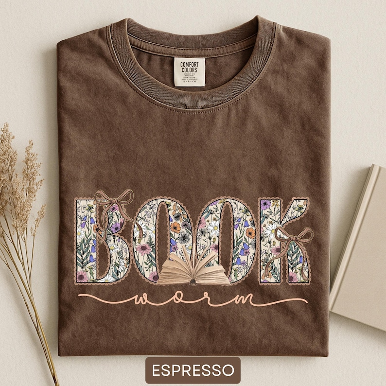 May include: Espresso-colored t-shirt with the words "BOOK worm" in a floral design. The word "BOOK" is filled with colorful flowers and an open book graphic. The shirt has a crew neck and a Comfort Colors tag.