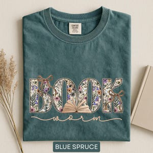 May include: Blue spruce t-shirt with the word "BOOK" in floral print, a small open book, and the word "worm" in a cursive font. The shirt has a crew neck and a tag that says "Comfort Colors".