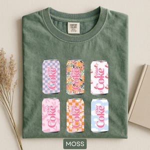May include: A moss green Comfort Colors t-shirt featuring six Diet Coke cans with various patterns, including floral, checkered, and cloud designs. The cans are in shades of pink, orange, and blue. The word "MOSS" is printed at the bottom of the shirt.