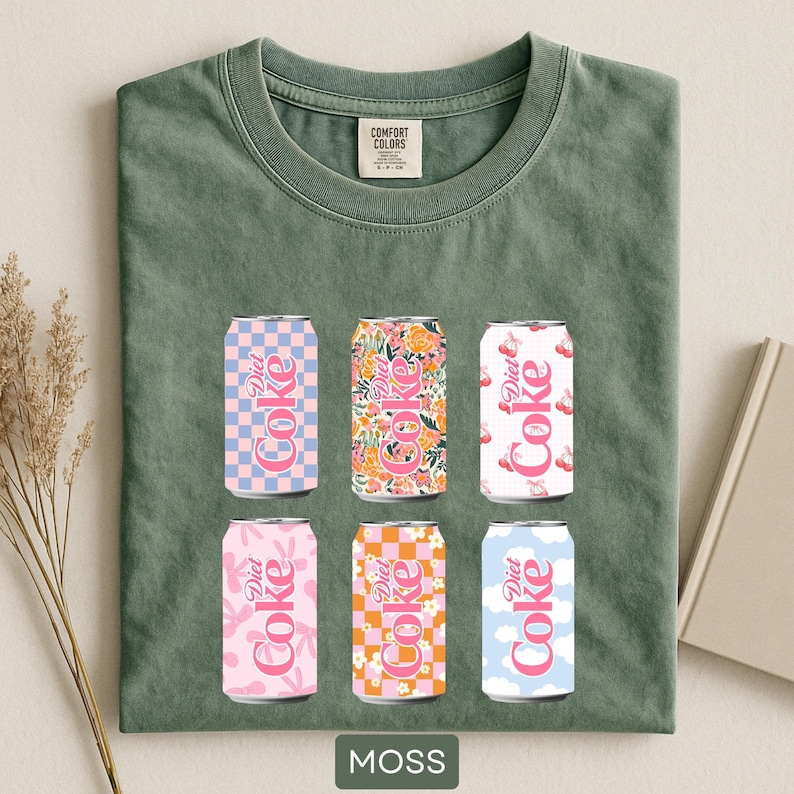 May include: A moss green Comfort Colors t-shirt featuring six Diet Coke cans with various patterns, including floral, checkered, and cloud designs. The cans are in shades of pink, orange, and blue. The word "MOSS" is printed at the bottom of the shirt.