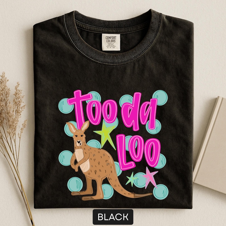 Op de afbeelding: Zwart T-shirt met een kangoeroe-afbeelding en de tekst "Tooda Loo" in roze. Het ontwerp bevat blauwe cirkels en groene en roze sterren. Het T-shirt is gemaakt van een zacht materiaal.