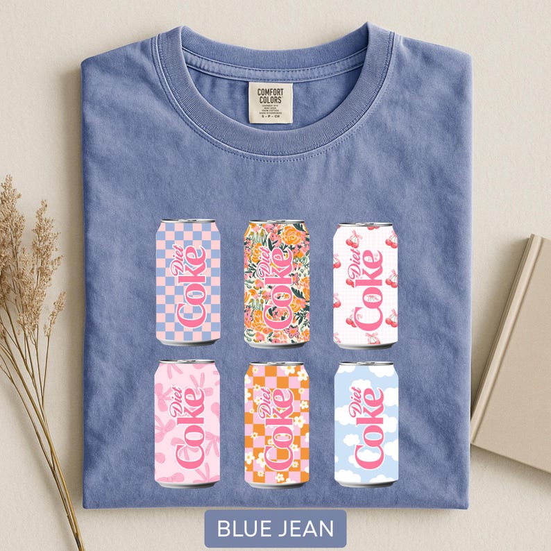 May include: A blue jean Comfort Colors t-shirt featuring six Diet Coke can designs. The cans have various patterns, including floral, checkered, and cloud motifs. The text "Diet Coke" is visible on each can. The shirt has a crew neck and a tag that says "Comfort Colors".