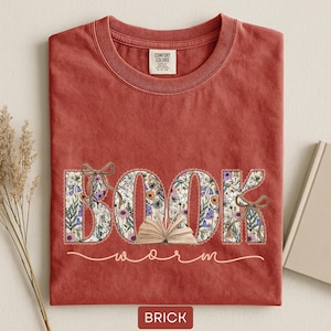 May include: Brick-colored t-shirt with a floral "BOOK" design and the word "worm" in a script font. The letters of "BOOK" are filled with a floral pattern and tied with a bow. A small open book is in the center.