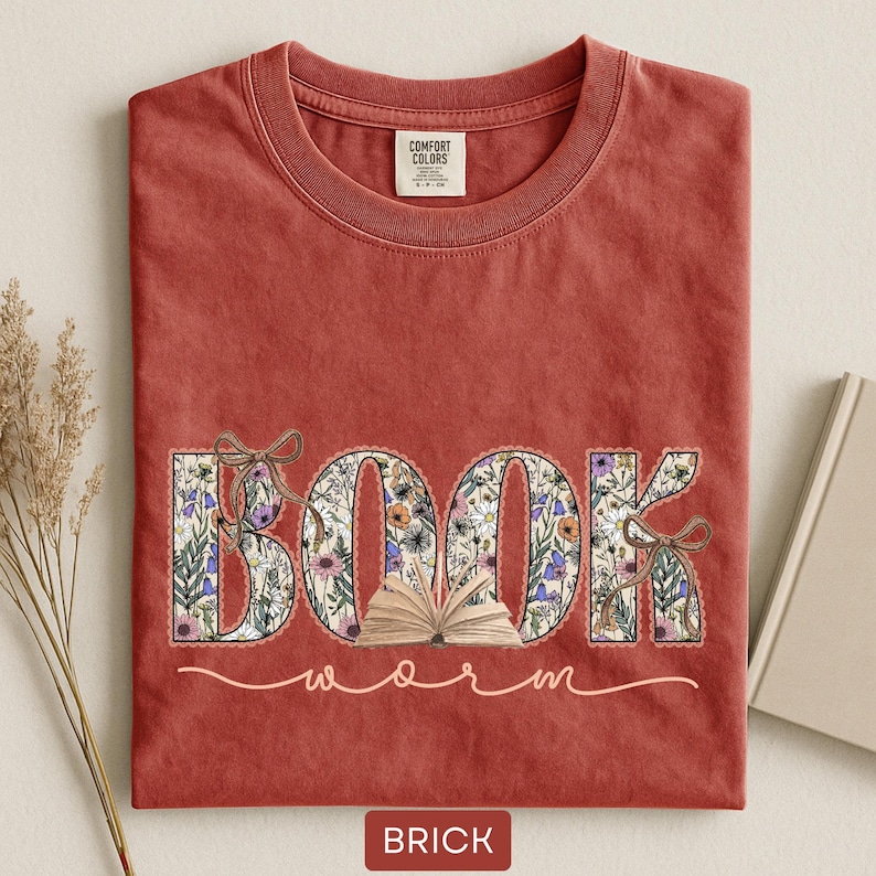 May include: Brick-colored t-shirt with a floral "BOOK" design and the word "worm" in a script font. The letters of "BOOK" are filled with a floral pattern and tied with a bow. A small open book is in the center.