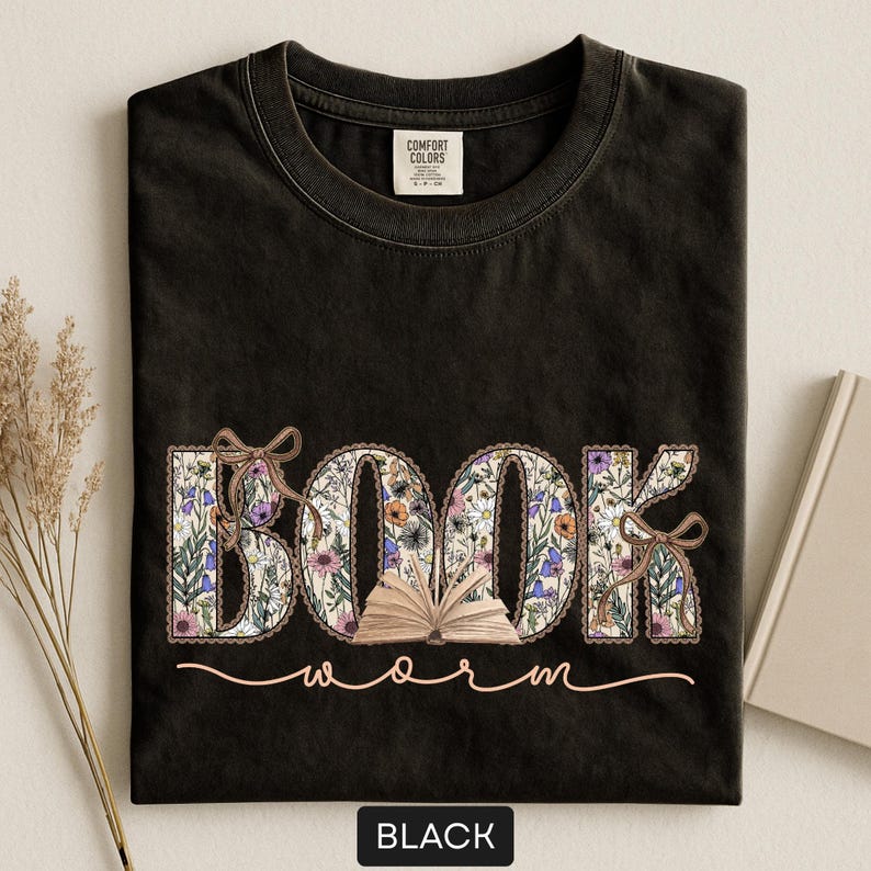 May include: Black t-shirt with the words "BOOK worm" in a floral design. The word "BOOK" is in large letters with a floral pattern, and the word "worm" is in a cursive font. A small open book is between the words.