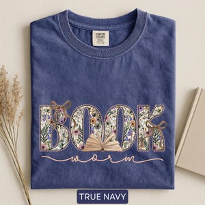 May include: A navy blue t-shirt with the words "BOOK worm" in a floral design. The word "BOOK" is filled with colorful flowers and tied with brown bows. The text "worm" is in a cursive font. The shirt has a Comfort Colors tag.