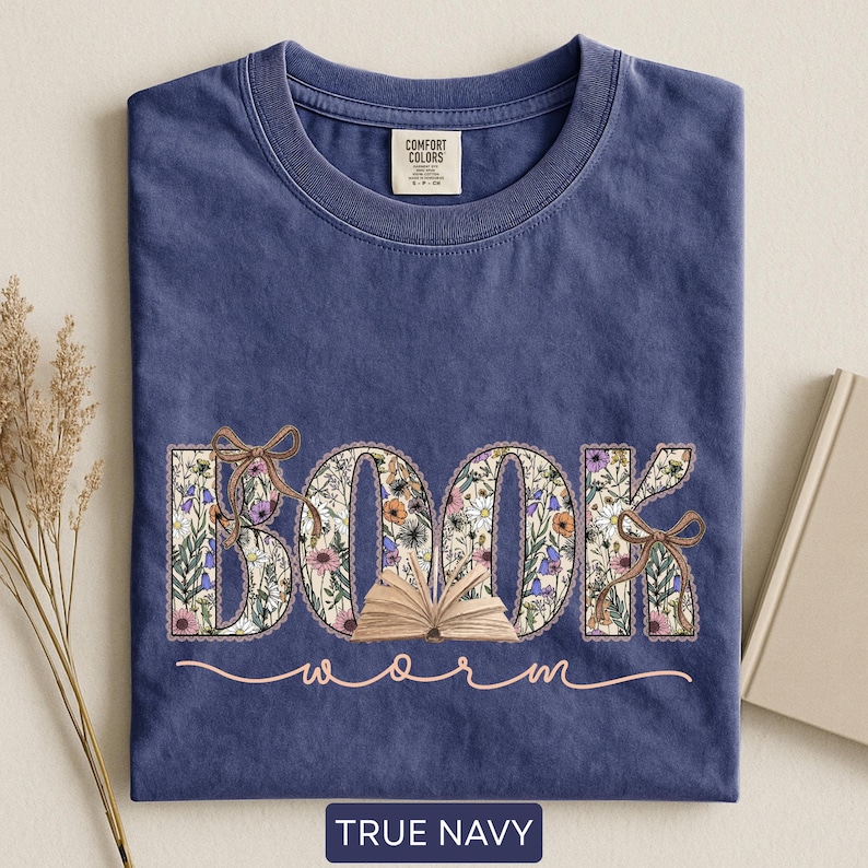 May include: A navy blue t-shirt with the words "BOOK worm" in a floral design. The word "BOOK" is filled with colorful flowers and tied with brown bows. The text "worm" is in a cursive font. The shirt has a Comfort Colors tag.