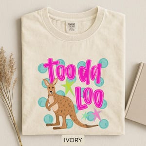 Op de afbeelding: Ivoor kleurig T-shirt met een kangoeroe illustratie en de tekst "Tooda Loo" in felroze. Het ontwerp bevat blauwe bellen en sterren accenten. De tekst "IVORY" staat onderaan.