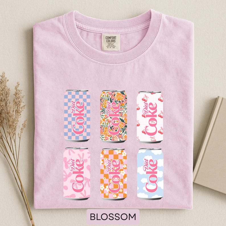 May include: A light pink t-shirt featuring six Diet Coke can designs with various patterns, including floral, checkered, and cloud motifs. The word "Blossom" is printed at the bottom. The shirt has a crew neck and short sleeves.