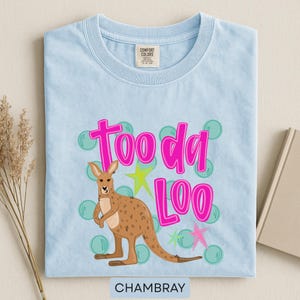 Op de afbeelding: Lichtblauw chambray T-shirt met een kangoeroe-afbeelding en de roze tekst "Tooda Loo". De kangoeroe is bruin en het ontwerp bevat groene bellen en gele en roze sterren. Het shirt heeft een ronde hals.