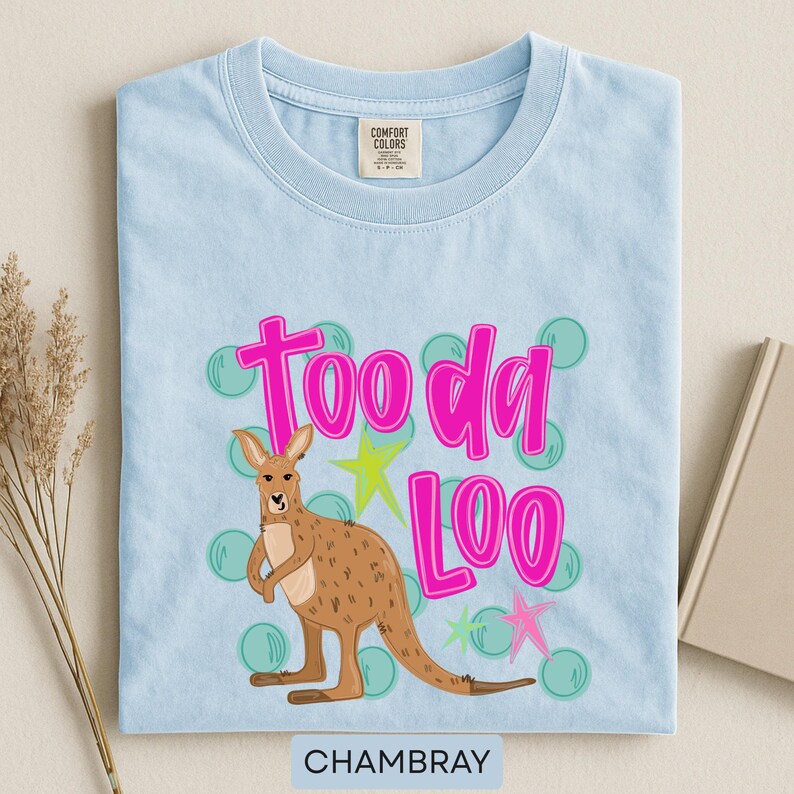Op de afbeelding: Lichtblauw chambray T-shirt met een kangoeroe-afbeelding en de roze tekst "Tooda Loo". De kangoeroe is bruin en het ontwerp bevat groene bellen en gele en roze sterren. Het shirt heeft een ronde hals.