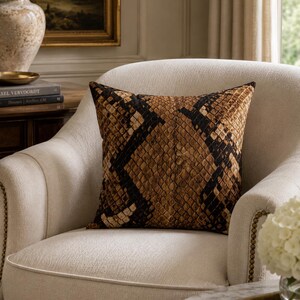 May include: A square throw pillow featuring a brown and black snakeskin pattern. The pillow has a textured surface with a repeating scale design in shades of tan, dark brown, and black, displayed on a cream-colored upholstered armchair.