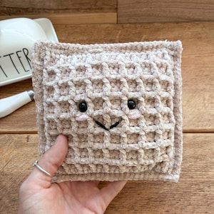 Puede incluir: Un juguete de peluche cuadrado de color beige con forma de gofre, hecho de hilo suave y texturizado. Presenta dos pequeños ojos redondos de plástico negro, una boca negra bordada en forma de V y dos pequeños círculos bordados en rosa claro.