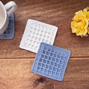 May include: Three square crochet coasters featuring a waffle stitch pattern. One coaster is white and two are light blue. The coasters are arranged with one blue coaster under a white ceramic mug and two others placed separately on a wooden surface.