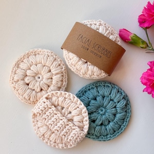 May include: Three round, crocheted facial scrubbies made of 100% cotton. Two scrubbies are cream-colored, and one is teal. A brown paper band is wrapped around the cream-colored scrubbies with the text FACIAL SCRUBBIES 100% COTTON printed on it.
