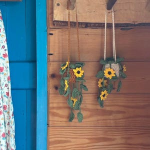 May include: Two handmade crochet plant hangers with sunflower and leaf details. One has a brown strap, the other a white strap. The hangers are suspended against a wooden wall. The text "PDF Pattern only" is visible at the bottom.