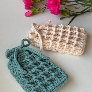 May include: Two handmade crochet bags in cream and teal, with a textured weave pattern. The cream bag has a drawstring closure, and the teal bag has a loop closure. Pink carnations are in the background.