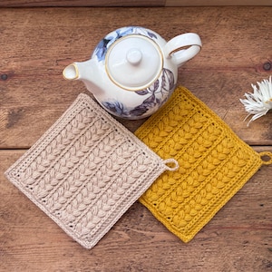 Op de afbeelding: Twee vierkante gehaakte pannenlappen, één in beige en één in mosterdgeel, met een gestructureerd vlechtpatroon en een klein lusje op de hoek. Een witte keramische theepot met een blauw en grijs bloemenpatroon staat boven de pannenlappen.