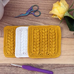 May include: Three square crochet motifs featuring a central braided cable stitch pattern. Two motifs are mustard yellow and one is white. The items are arranged horizontally alongside a purple crochet hook and iridescent stork-shaped scissors.