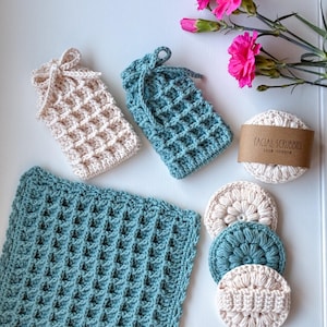 May include: A set of crochet items including a large teal square washcloth, two small rectangular pouches in cream and teal with drawstring ties, and three round facial scrubbies in cream and teal, with a paper band labeled FACIAL SCRUBBIES.