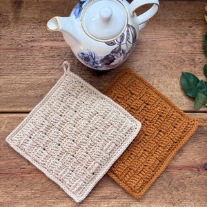 Könnte beinhalten: Eine weiße Teekanne aus Porzellan mit blauem Blumenmuster und Goldrand, begleitet von zwei quadratischen Häkel-Topflappen. Ein Topflappen ist hellbeige und der andere senfgelb, beide mit einem strukturierten Korbgeflecht-Muster.