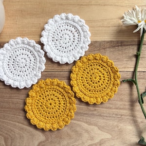 May include: Four circular crochet coasters arranged in a cluster, featuring two white and two mustard yellow pieces. Each coaster has a textured center and a scalloped, bobble-stitch border. A white daisy flower is positioned to the right side.