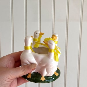 May include: A white ceramic planter featuring four sculpted ducks standing in a circle on a green base. Each duck has a yellow ribbon tied around its neck. The ducks have orange beaks and feet, and the centre of the planter is hollow.