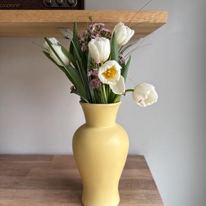 May include: A yellow vase holding a bouquet of white tulips with green leaves and small purple flowers. The vase sits on a wooden surface. A vintage radio is on a wooden shelf in the background.