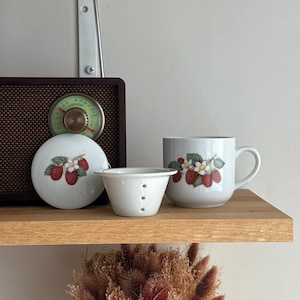 Peut inclure: Un mug en céramique blanche et son couvercle circulaire assorti, tous deux ornés d'une illustration botanique de framboises rouges, de feuilles vertes et de fleurs blanches. Une petite passoire à thé en céramique blanche est placée devant.