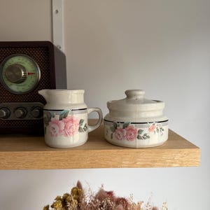 Peut inclure: Ensemble de pot à crème et sucrier en céramique crème. Chaque pièce présente un motif floral avec des roses roses et des feuilles vertes, accentué de rayures noires. L'ensemble est présenté sur une étagère en bois.
