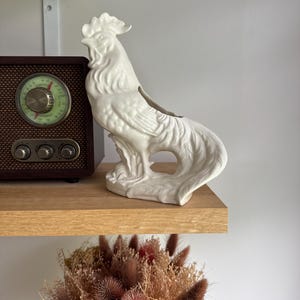 May include: A white ceramic rooster vase sits on a wooden shelf next to a vintage radio. The rooster has detailed feather textures and a hollowed-out back for displaying flowers. A bouquet of dried flowers is below the shelf.