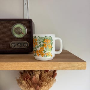 Peut inclure: Une tasse en verre opalin blanc ornée d'un motif répétitif de fleurs orange vif et de feuilles vertes. La tasse présente un corps cylindrique avec une anse en forme de C et une base légèrement encastrée, arborant un imprimé botanique éclatant.