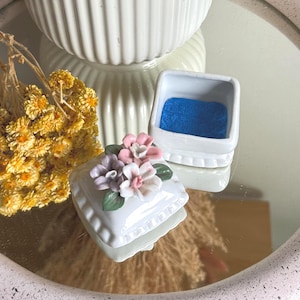 May include: A small white ceramic trinket box with a square base and a removable lid. The lid is decorated with three sculpted ceramic flowers in shades of pink, lavender, and white with green leaves. The interior of the box is lined with blue velvet.