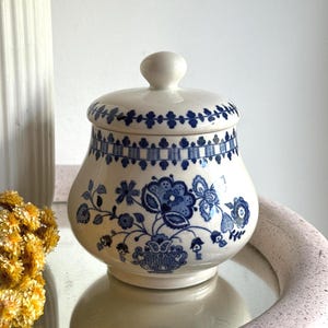 Peut inclure: Un sucrier en céramique de couleur crème avec un couvercle assorti, orné d'un motif floral bleu et d'une bordure décorative sur le dessus. Le bol présente un corps arrondi et un petit bouton sur le couvercle, avec des motifs botaniques bleus.