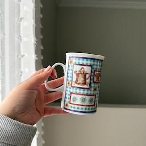 Peut inclure: Une tasse en céramique blanche avec un motif à carreaux bleu et blanc et des illustrations florales et d'arrosoirs. La tasse a une anse incurvée et un bord blanc.