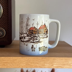 May include: A white ceramic mug featuring a detailed brown ink illustration of a harbour scene with sailboats, wooden buildings with orange roofs, and birds. The base of the mug has a soft blue gradient fade, and it includes a sturdy handle.