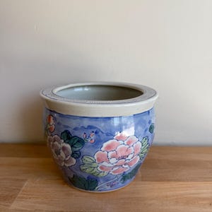 Op de afbeelding: Een ronde, blauwe keramische plantenbak met een witte rand en bloemenmotief. De plantenbak heeft roze en paarse bloemen met groene bladeren tegen een blauwe achtergrond. De rand heeft een decoratief patroon.