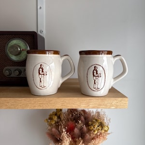 May include: A white ceramic mug with a black handle and the text "But First, Coffee" in a bold, black font. The mug is set against a blurred background, suggesting a cozy setting.