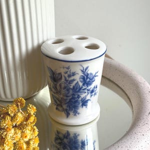 May include: A white ceramic toothbrush holder featuring a blue floral pattern and a blue rim. The top of the holder has four circular openings for storage. The cylindrical body tapers slightly towards the base, showcasing a detailed botanical design.