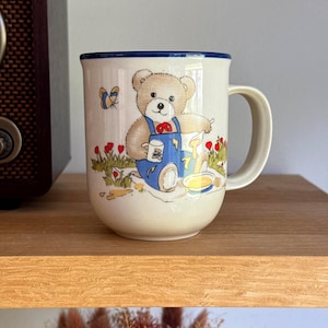 May include: A cream-coloured ceramic mug with a dark blue rim and a handle. The front features a graphic of a brown teddy bear wearing blue overalls, sitting in a field of red flowers, holding a small white cup and pouring yellow liquid into a bowl.