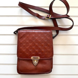 May include: A brown leather crossbody bag featuring a woven texture on the front flap, a gold-tone metal turn-lock closure, and an adjustable shoulder strap with a gold-tone buckle. The bag has a smooth leather lower section and a structured shape.