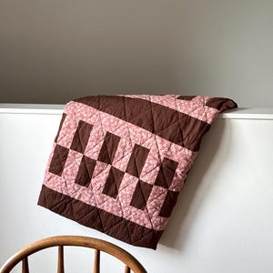 May include: A quilted blanket featuring a geometric pattern of solid dark brown rectangular blocks alternating with light pink floral patterned blocks. The fabric has a diamond-shaped stitched texture throughout the entire surface of the quilt.