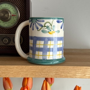 Peut inclure: Une tasse en céramique avec une anse et une base blanches. La tasse présente un motif à carreaux bleus et jaunes sur la moitié inférieure et des motifs floraux sur la moitié supérieure. Le bord et la base sont verts.